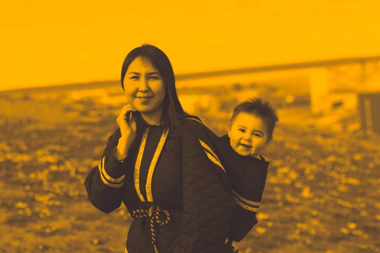 An Inuk woman wearing a decorated amauti, a traditional Inuit mother’s parka, smiles at the camera, as does her baby who peers out of the hood of the amauti which hangs off her back.