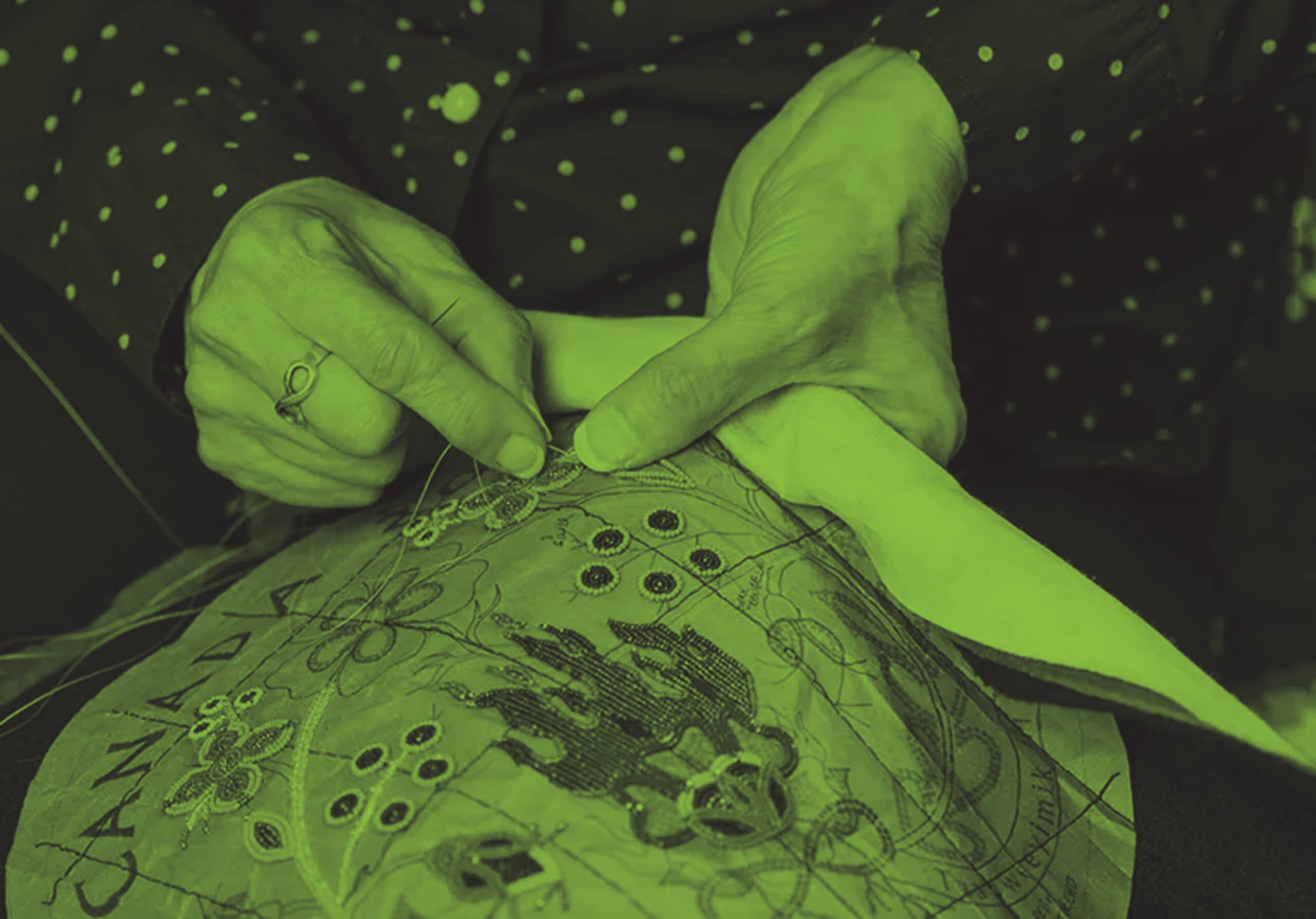 Les mains de l’artiste métisse Jennine Krauchi tiennent un morceau de tissu brodé sur lequel elle ajoute une broderie perlée à motifs floraux autour d’un motif représentant un feu. Le symbole de l’infini métis et le mot « Canada » sont visibles sur le tissu.