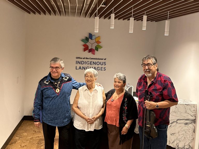 4 Indigenous Elders standing together in front of a white wall with the Commission's logo behind them.